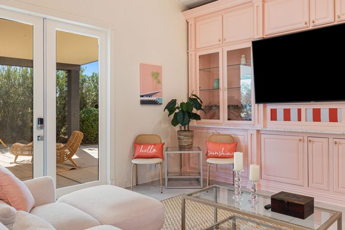 Sunlit modern living room with blush-pink built-in cabinets and mounted TV, glass coffee table with candles, two rattan chairs with coral "hello" and "sunshine" pillows, potted plant and sliding glass doors to a patio with wicker loungers.
