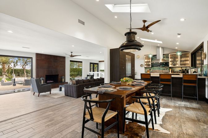 Open-concept modern dining and living area with a rustic wood table set for six, black woven-seat chairs, pendant lights and skylight, dark-wood kitchen island with barstools, fireplace wall, large sliding glass doors to a patio and trees, wood-look tile floors.