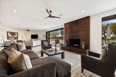 Cozy modern open-plan living room with dark sectional sofa, accent chairs and coffee table, large wood-paneled fireplace, ceiling fans, and sliding glass doors opening to a sunny patio with a striped umbrella and trees.