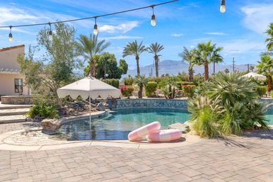 Sunny desert backyard pool with palm trees, string lights, scalloped umbrella and two pink inflatable rings on a stone deck, mountains in the distance.