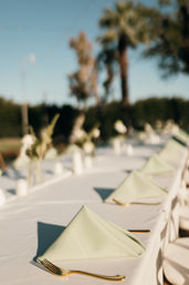 Sunlit outdoor banquet table set for an event with folded sage-green napkins, gold flatware, white tablecloths and blurred palm trees in the background