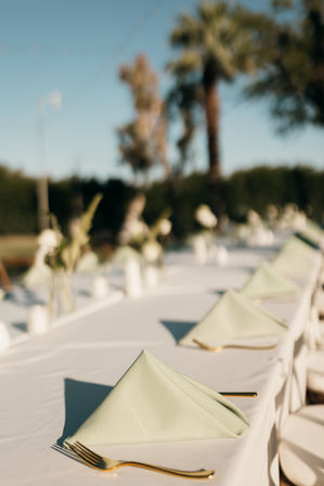 Sunlit outdoor banquet table set for an event with folded sage-green napkins, gold flatware, white tablecloths and blurred palm trees in the background