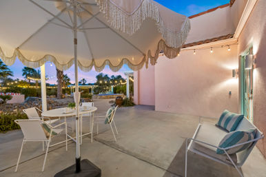 Twilight outdoor patio with a fringed white umbrella, white metal dining set and bench with teal-striped cushions, string lights over a palm-lined yard and pool, pink stucco wall and mint-green door