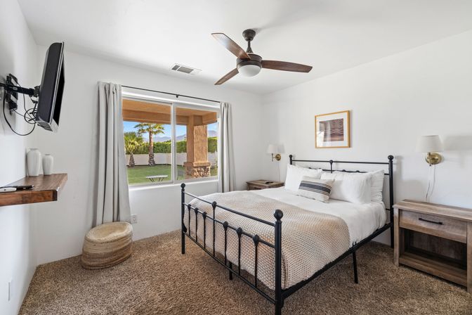 Cozy sunlit bedroom with black metal bed, knit throw and wood nightstands, ceiling fan and wall-mounted TV; large window overlooking covered patio, lawn, palm trees and distant mountains.