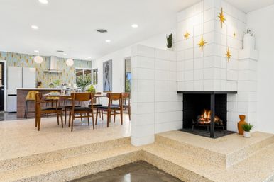 Mid-century modern open-plan dining and kitchen with terrazzo steps, wooden dining set and round table, kitchen island with pendant lights and colorful geometric tile backsplash, white block fireplace with burning fire and gold starburst wall decor.