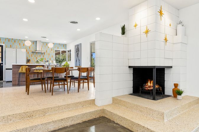 Mid-century modern open-plan dining and kitchen with terrazzo steps, wooden dining set and round table, kitchen island with pendant lights and colorful geometric tile backsplash, white block fireplace with burning fire and gold starburst wall decor.