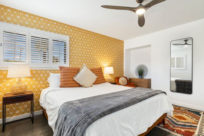 Bright modern bedroom with mustard-yellow geometric accent wall, large bed with gray throw and rust pillows, shuttered windows, rattan bedside lamps, ceiling fan, full-length mirror and patterned rug.