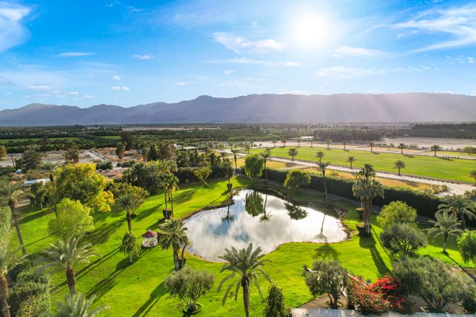 Aerial view of a sunlit desert oasis with a reflective pond, manicured green lawns, palm trees and a small gazebo, framed by distant mountains and a bright blue sky.