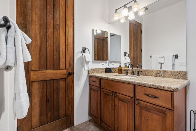 Cozy residential bathroom with rustic wooden door, granite countertop vanity, warm wood cabinets, oil-rubbed bronze faucets, large mirror and white towels