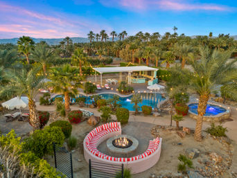 Aerial view of a desert oasis-style backyard at twilight with palm trees, mountains, a lagoon-shaped pool lit by string lights, a round red-and-white cushioned fire pit, and a glowing hot tub.
