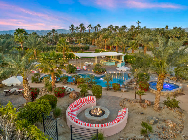 Aerial view of a desert oasis-style backyard at twilight with palm trees, mountains, a lagoon-shaped pool lit by string lights, a round red-and-white cushioned fire pit, and a glowing hot tub.