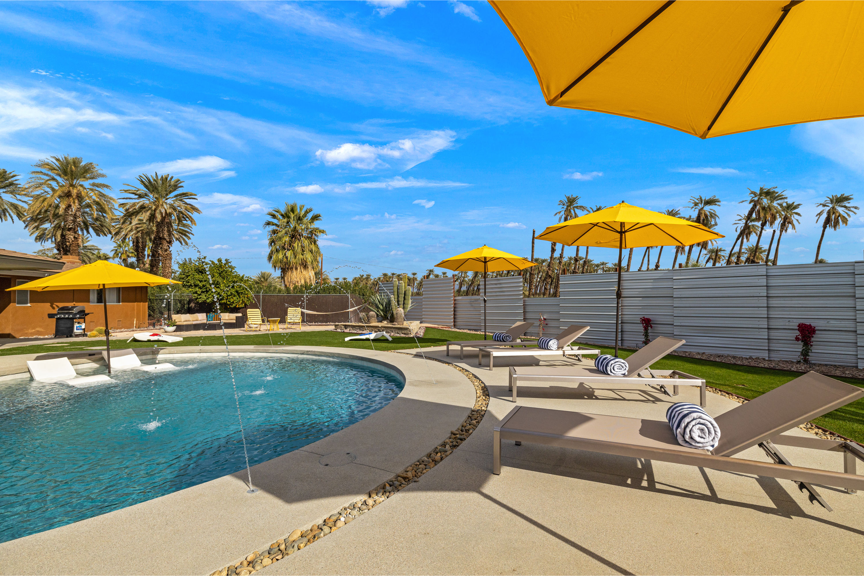 Sunlit backyard pool with water fountains, palm trees and bright yellow umbrellas shading lounge chairs with rolled towels under a clear blue sky — desert oasis vibe.