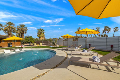 Sunlit backyard pool with water fountains, palm trees and bright yellow umbrellas shading lounge chairs with rolled towels under a clear blue sky — desert oasis vibe.
