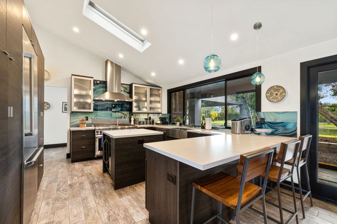 Modern open-concept kitchen with white quartz island and breakfast bar, dark wood cabinets, stainless steel appliances, blue glass pendant lights and skylight, wood-look tile floors and large sliding window overlooking a lush backyard patio.