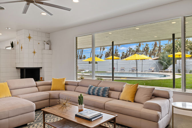 Bright midcentury-modern living room with a beige sectional and yellow pillows, wood coffee table with a small cactus, white tiled fireplace with gold star accents, and floor-to-ceiling windows looking out to a backyard pool with yellow umbrellas and palm trees.
