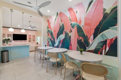Tropical-chic pastel dining area with a large pink and green banana-leaf mural, round white tables and rattan chairs, mint-blue service counter with pendant lights, ceiling fans and tiled floor