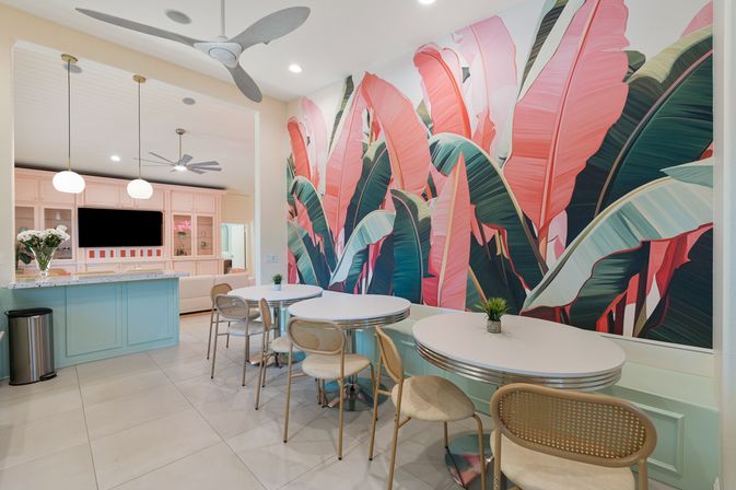 Tropical-chic pastel dining area with a large pink and green banana-leaf mural, round white tables and rattan chairs, mint-blue service counter with pendant lights, ceiling fans and tiled floor
