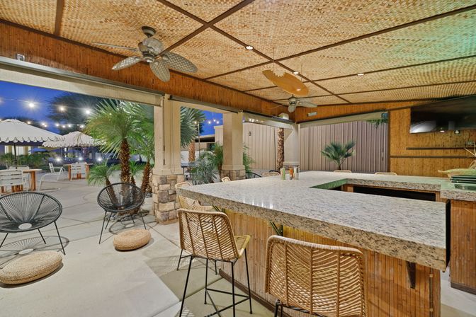 Tropical-inspired outdoor patio bar at dusk with a granite L-shaped counter, rattan bar stools, woven bamboo ceiling, ceiling fans, string lights and palm plants.