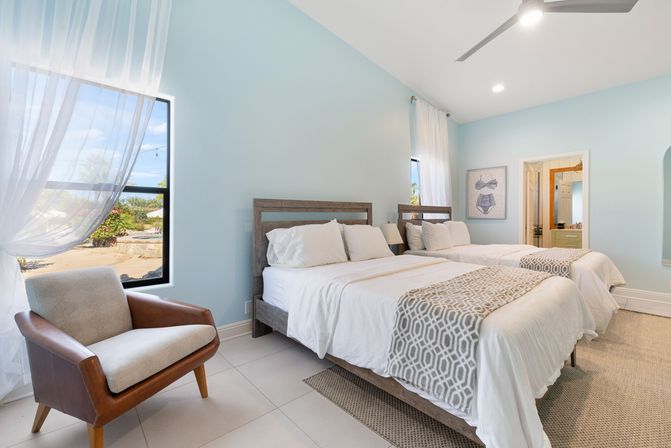 Sunny coastal-style guest bedroom with two twin beds in white linens, geometric throws, wooden headboards, leather accent chair by a sheer-curtained window overlooking tropical landscaping.