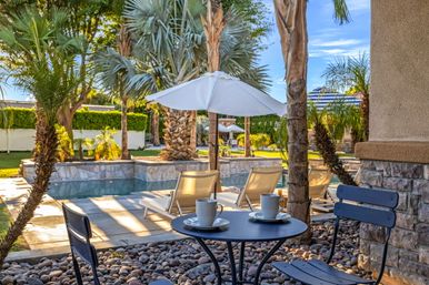 Sunny tropical-style backyard pool patio with palm trees, lounge chairs under a white umbrella and a bistro table holding two coffee cups