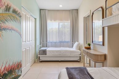 Coastal-inspired sunlit guest bedroom with white metal daybed, tropical palm mural, neutral curtains and wooden console with mirror
