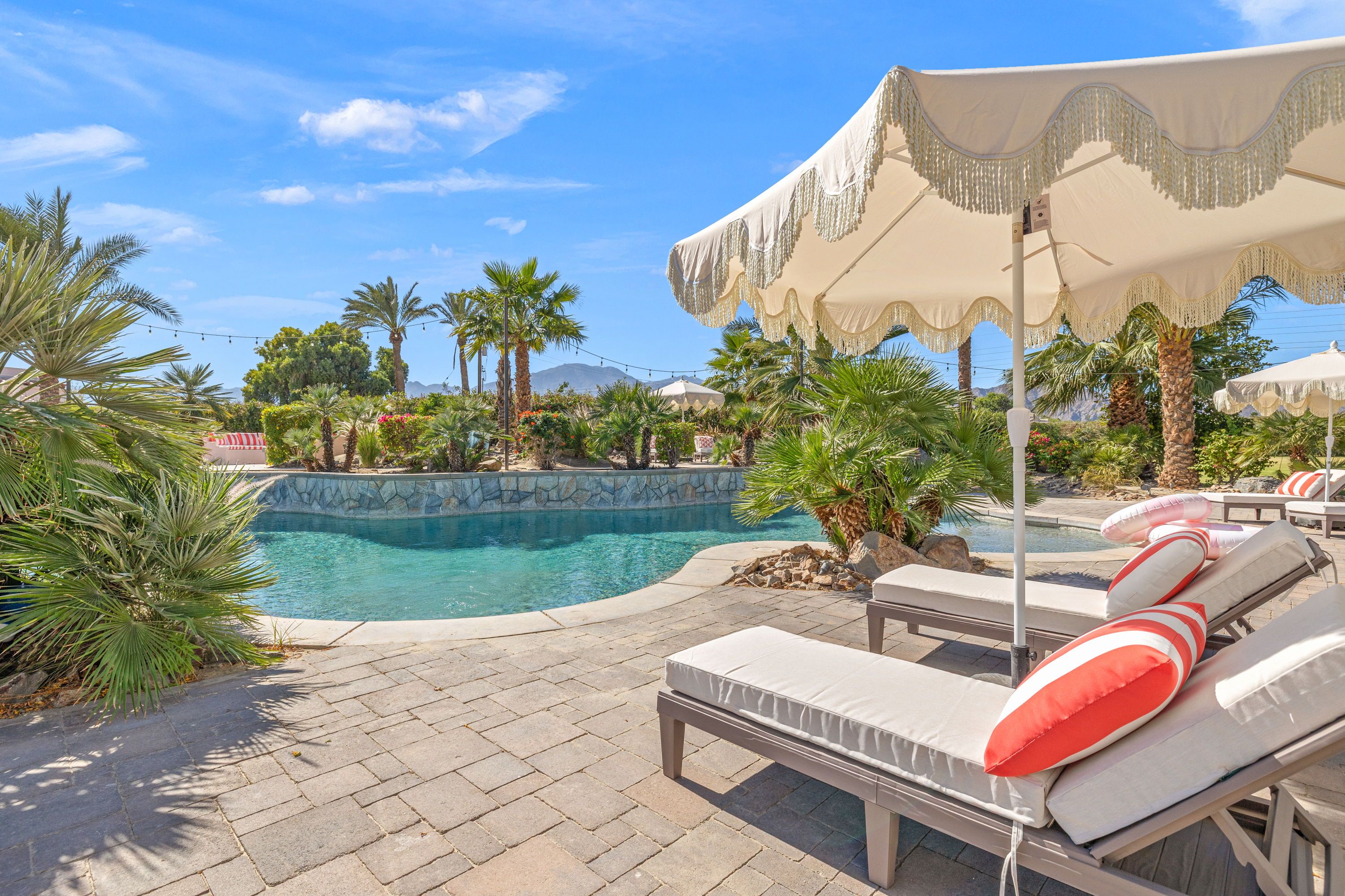 Sunny desert-oasis pool with turquoise water, palm trees, fringed cream parasols and cushioned lounge chairs topped with coral-striped pillows on a paver deck, mountains in the background.