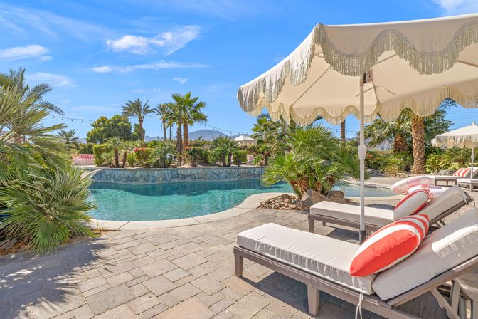 Sunny desert-oasis pool with turquoise water, palm trees, fringed cream parasols and cushioned lounge chairs topped with coral-striped pillows on a paver deck, mountains in the background.