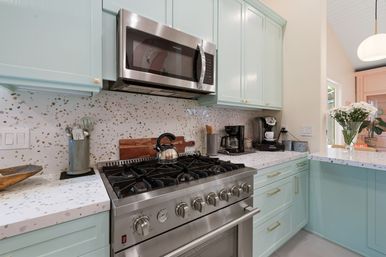Cheerful kitchen with mint-green cabinets, terrazzo countertops and backsplash, stainless-steel gas range and microwave, coffee station and vase of white daisies.