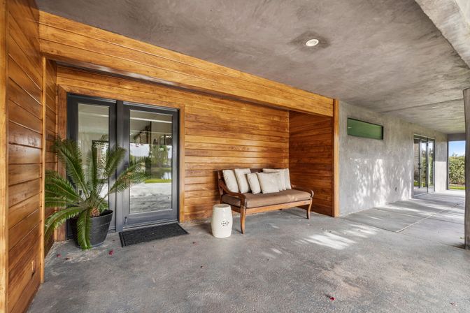 Covered modern patio entry with warm horizontal wood siding, glass double doors, concrete floor, potted palm, and a cushioned wooden bench — cozy outdoor seating nook