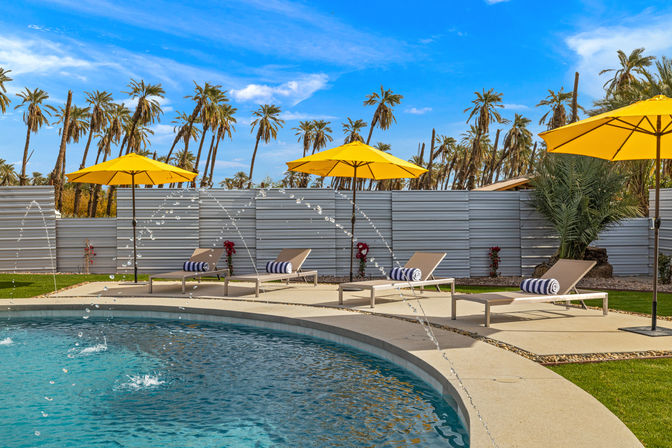 Sunny outdoor pool with arcing water jets, beige lounge chairs topped with navy-and-white striped towels, bright yellow umbrellas and tall palm trees under a vivid blue sky.