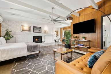 Mid-century modern open-plan studio with white brick fireplace, wood-paneled TV wall, leather sofa, bed, geometric rug and large indoor plants.