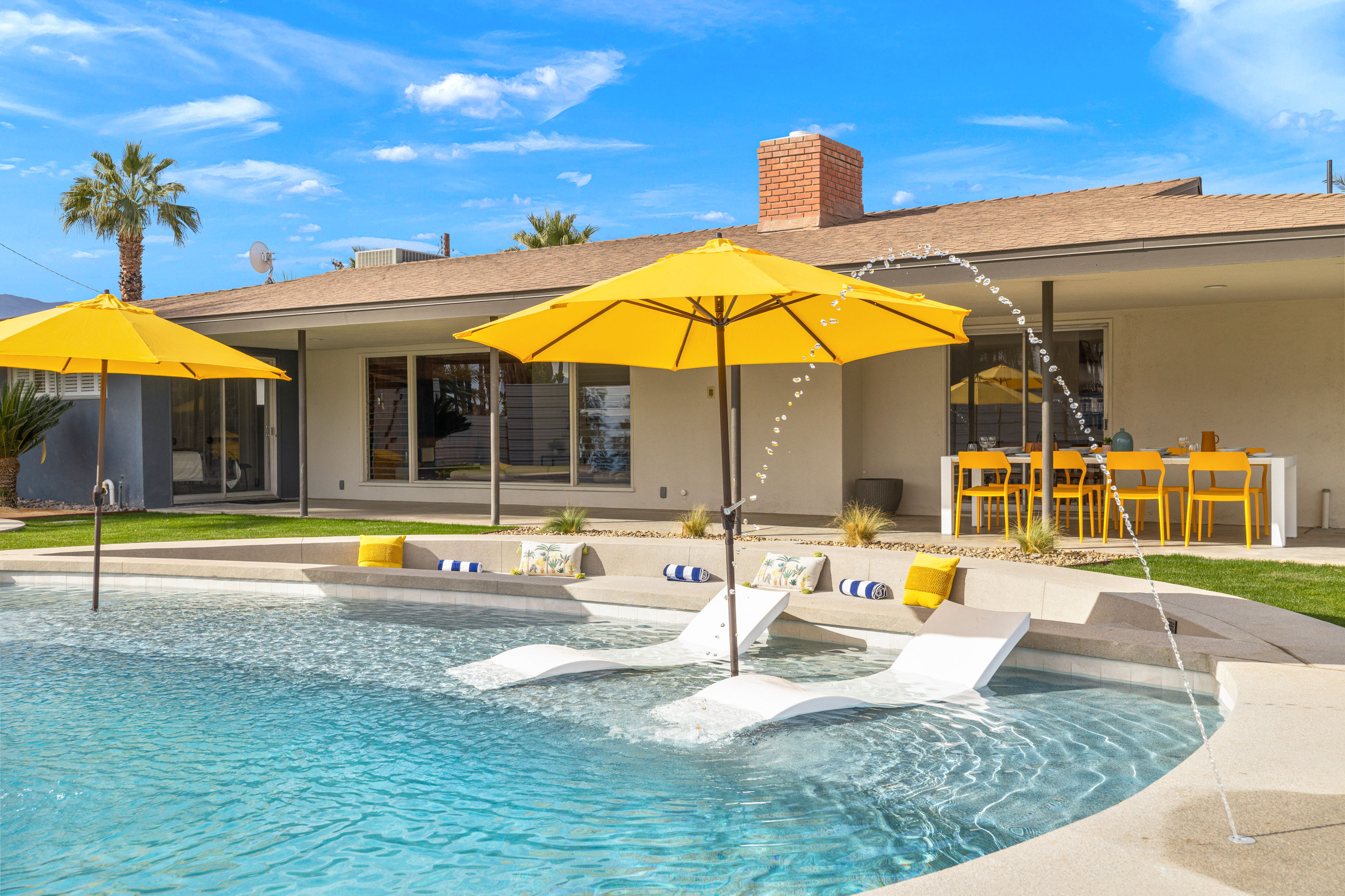 Sunny backyard pool with yellow umbrellas and in-pool loungers, arcing water jets, palm trees, and a bright outdoor dining area beside a modern single-story home.