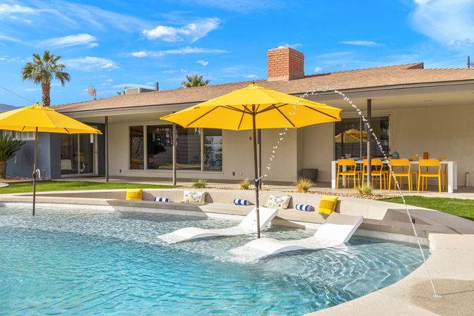 Sunny backyard pool with yellow umbrellas and in-pool loungers, arcing water jets, palm trees, and a bright outdoor dining area beside a modern single-story home.