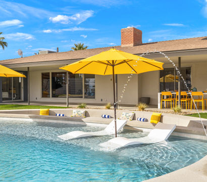 Sunny backyard pool with yellow umbrellas and in-pool loungers, arcing water jets, palm trees, and a bright outdoor dining area beside a modern single-story home.