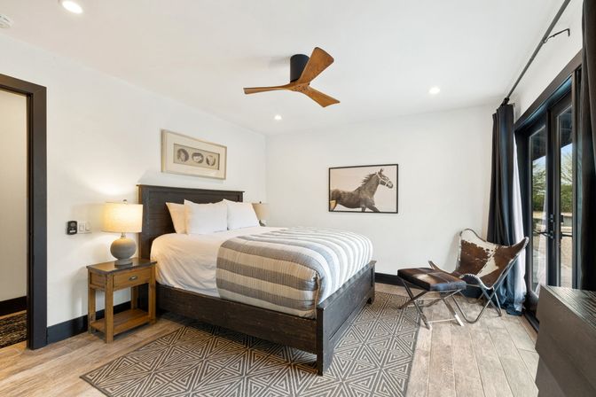 Cozy modern-rustic bedroom with dark wood king bed and striped duvet, geometric rug, wooden ceiling fan, bedside table with lamp, cowhide butterfly chair and floor-to-ceiling glass doors letting in natural light.