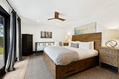 Bright modern bedroom with wooden king bed and headboard, patterned rug, black metal daybed, matching nightstands, ceiling fan and sliding glass door