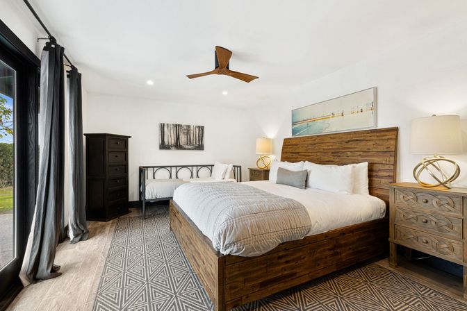 Bright modern bedroom with wooden king bed and headboard, patterned rug, black metal daybed, matching nightstands, ceiling fan and sliding glass door