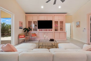 Sunlit coastal living room with pastel pink built-in media wall and mounted TV, plush white boucle sectional, glass coffee table on a woven rug, coral accent chairs with 'hello' and 'sunshine' pillows, and a sliding door opening to a patio garden.