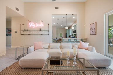 Sunlit open-concept living room with oversized cream sectional and pink throw pillows, glass coffee table with candles and remote on a woven rug, barstools and globe pendant lights at the kitchen island, and a pink neon "Stay Locale" sign on a wall-mounted glass shelf.