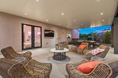 Covered suburban patio at dusk with rattan lounge chairs and orange accent pillows around a round coffee table, outdoor dining table, wall-mounted TV, and string lights over a landscaped backyard.