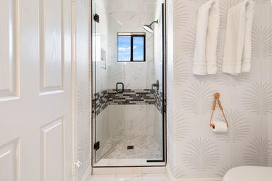 Modern home bathroom with glass walk-in shower, marble-look tiles and black mosaic accent band, rain shower head, white towels on wall hooks and a toilet paper roll on a rope holder