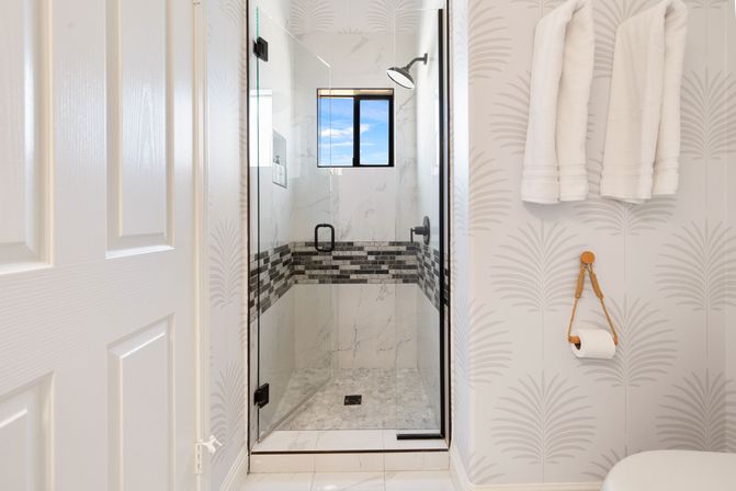 Modern home bathroom with glass walk-in shower, marble-look tiles and black mosaic accent band, rain shower head, white towels on wall hooks and a toilet paper roll on a rope holder