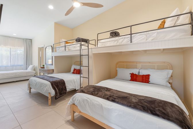 Bright family bunk bedroom in a vacation rental with two lower beds and two upper bunks, white linens, red accent pillows, metal ladder, ceiling fan and large sunlit window