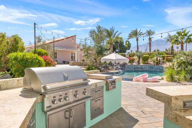 Sunny desert oasis backyard with built-in stainless steel grill and stone counters, curved pool with pink inflatable rings, umbrella, string lights, palm trees and mountain backdrop