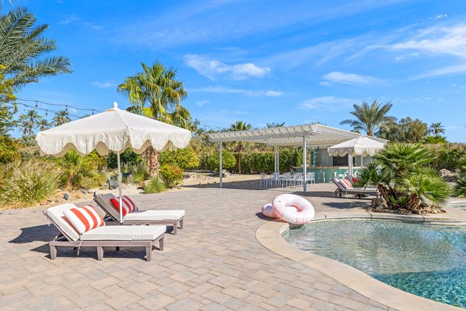 Sunny resort-style outdoor pool deck with white fringe parasols, cushioned sun loungers with red-striped pillows, pink inflatable rings by the curved pool, a pergola-covered poolside bar, and palm trees under a bright blue sky.