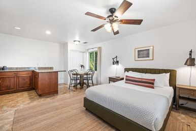 Bright studio apartment bedroom with king bed and olive headboard, granite-counter kitchenette with wooden cabinets, small round dining nook, ceiling fan and window view of greenery