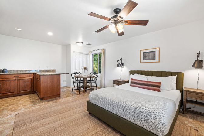 Bright studio apartment bedroom with king bed and olive headboard, granite-counter kitchenette with wooden cabinets, small round dining nook, ceiling fan and window view of greenery