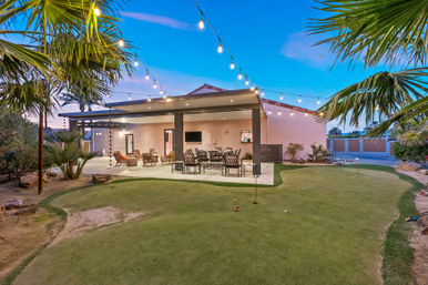 Inviting dusk backyard patio with covered dining and lounge area, string lights, palm trees and a small putting green in a desert-style residential yard.