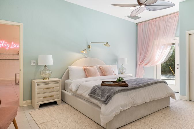 Light-filled pastel bedroom with mint walls, upholstered king bed in white linens, blush pillows, glass bedside lamps, brass swing sconce, leaf‑blade ceiling fan, sheer pink curtains tied by patio doors and a tray with a small plant on the bed.