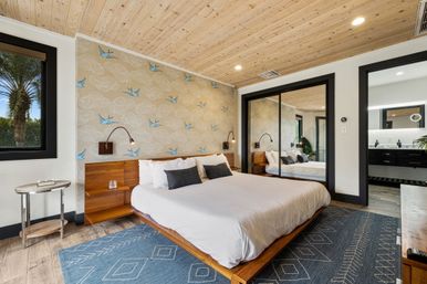 Modern coastal master bedroom with wood-paneled ceiling, king platform bed with white linens, blue-bird patterned accent wall, mirrored sliding closet, bedside reading lamps, blue geometric rug and a palm-tree view through the window.